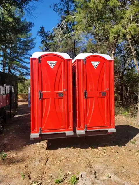 Two red PrimeTime portable toilets stamped with the triangle logo under pine trees