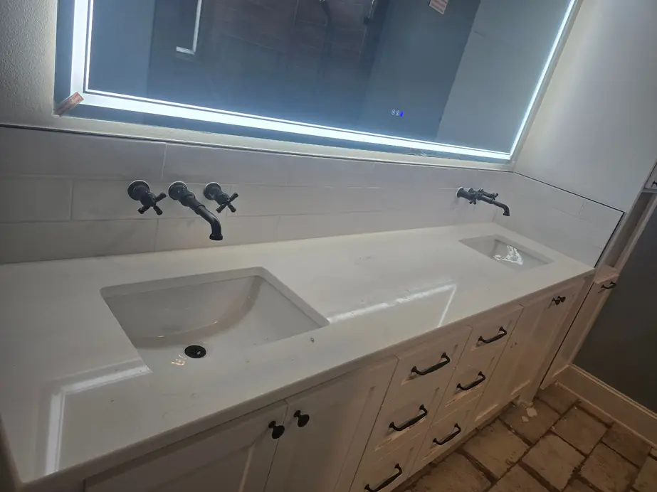 Double vanity with wall-mounted black faucets under a lit mirror, plumbed by PrimeTime